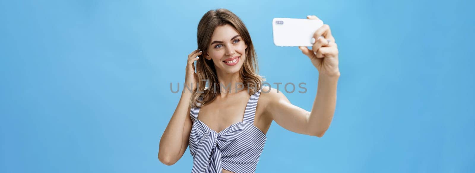 Attractive woman with good self-esteem in stylish matching outfit flicking hair behind ear smiling joyfully at smartphone screen, taking selfie to post in internet and gain followers over blue wall. Lifestyle concept