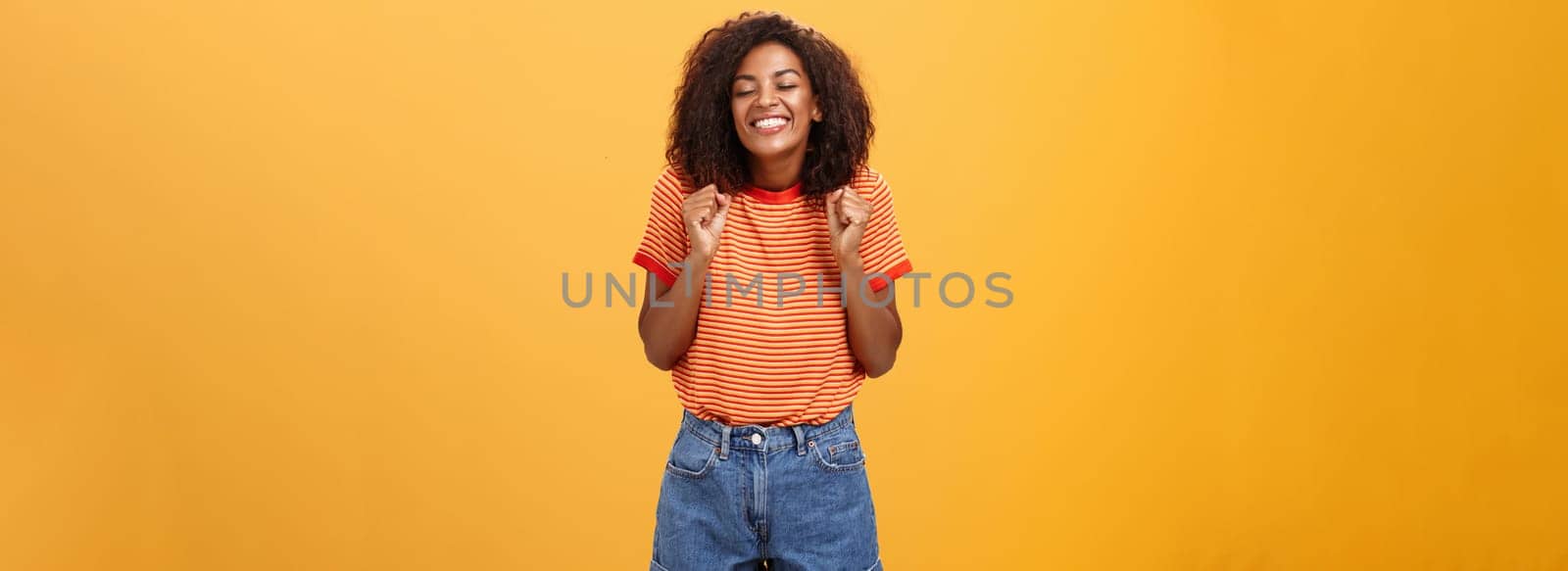 Girl eager to see lover coming from trip standing excited and thrilled with happy broad smile clenching fists grinning with closed eyes feeling joyful and amused over orange background by Benzoix