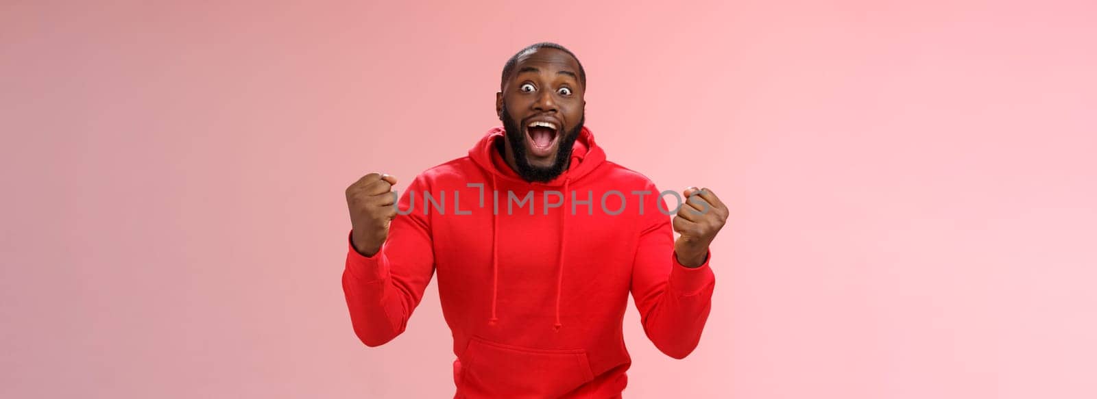 Very happy lucky black bearded guy winning lottery made winning bet clench fists triumphing yelling yes widen eyes surprised joyfully celebrating good news standing impressed, pink background.