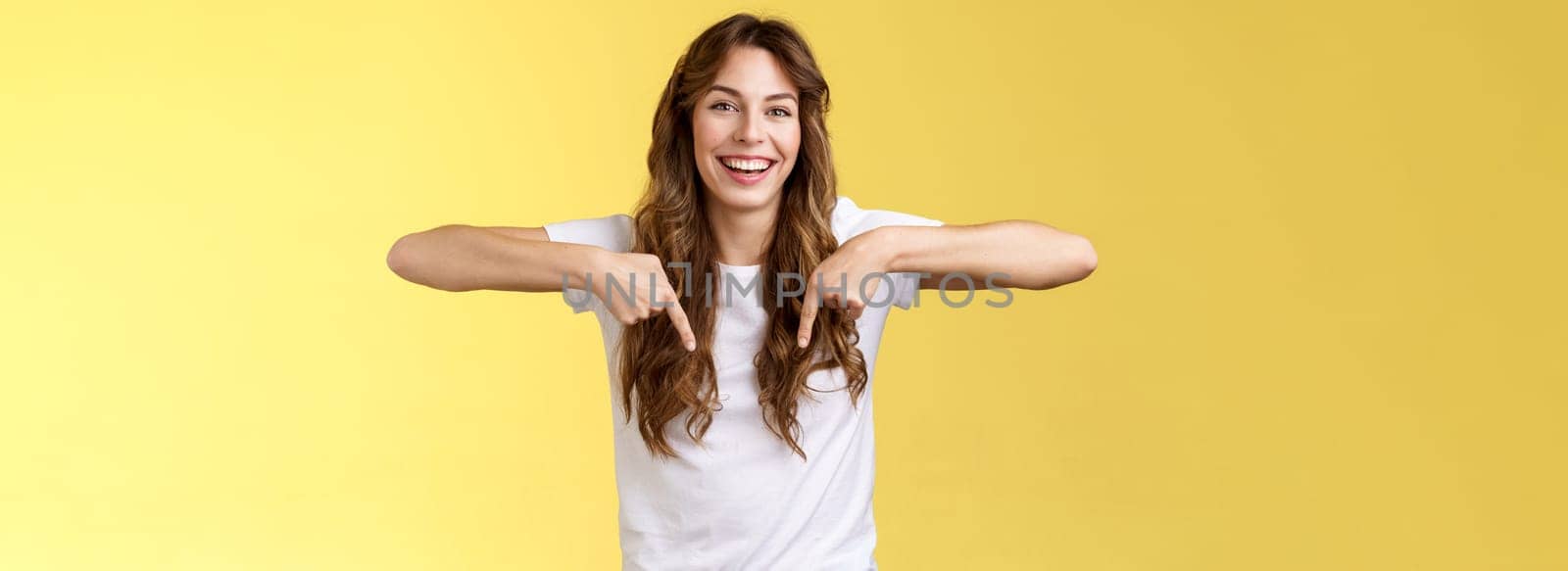 Cheerful lively happy funny girl long beautiful hair pointing down index fingers indicate bottom promo laughing joyfully share positive emotions recommend product advertising yellow background by Benzoix