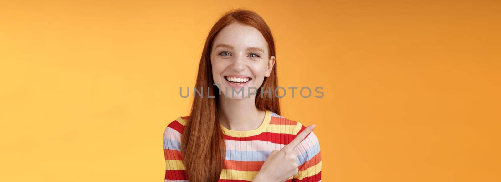 Happy charismatic smiling young ginger girl blue eyes sincere tender grin laughing broadly having fun discuss hilarious concert pointing upper left corner showing awesome product, orange background.