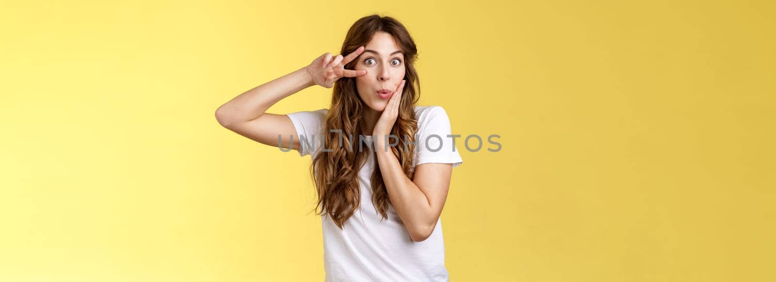 Silly coquesttish good-looking caucasian girl curly hairstyle folding lips amused interested touch cheek feminine sensual pose show victory peace sign upbeat stand intrigued yellow background by Benzoix