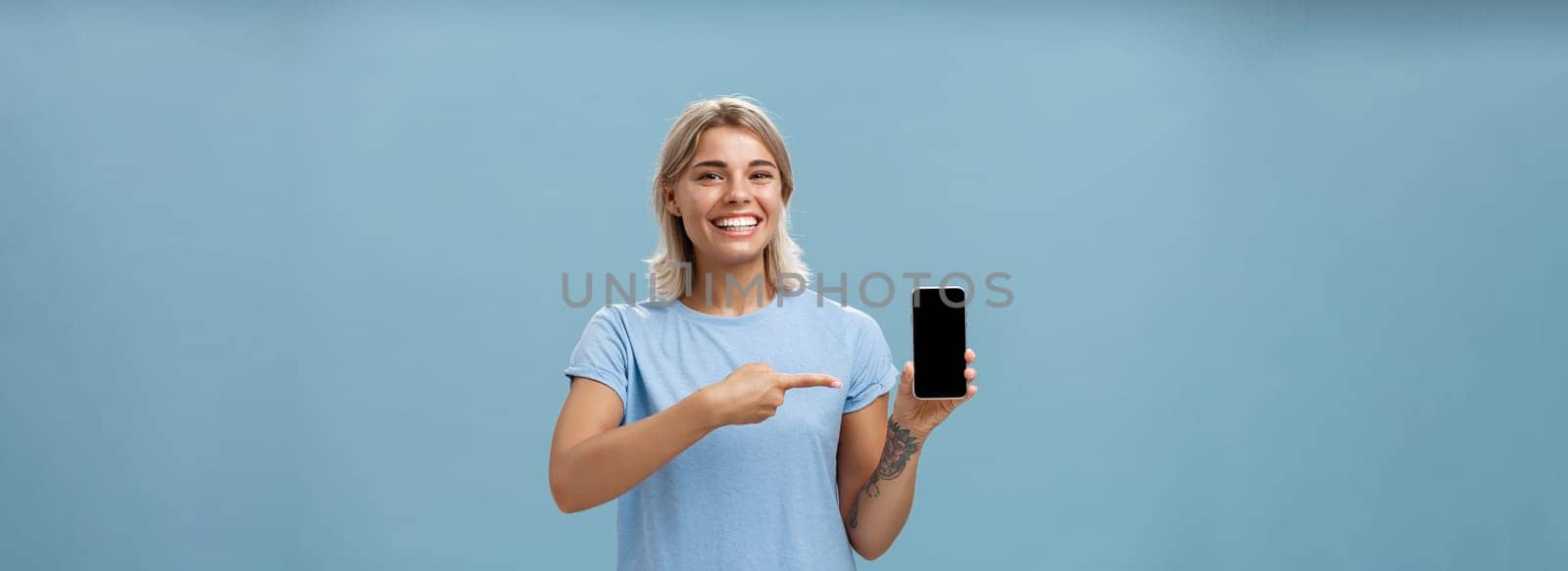 Cute enrgized girl showing smartphone. Charming blonde young female with cool tattoos laughing and smiling from happiness and joy pointing at device holding phone with screen faced to camera.