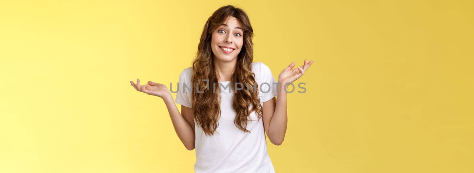 Who cares have fun. Carefree indifferent outgoing happy young woman shrugs raise hands sideways clueless unbothered apathic to topic uninterested smiling broadly careless yellow background.