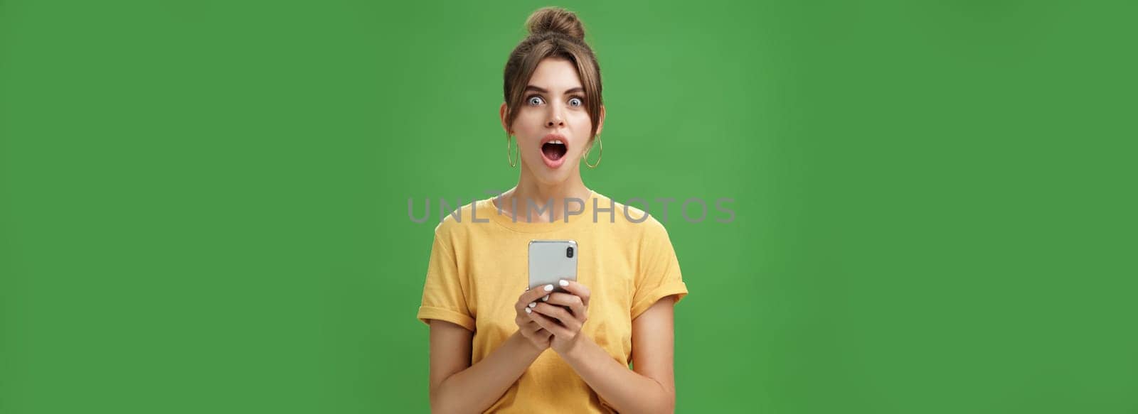 Portrait of shocked speechless and impressed beautiful white girl with combed hair in yellow t-shirt holding smartphone, dropping jaw from excitement reacting to cool app over green background by Benzoix