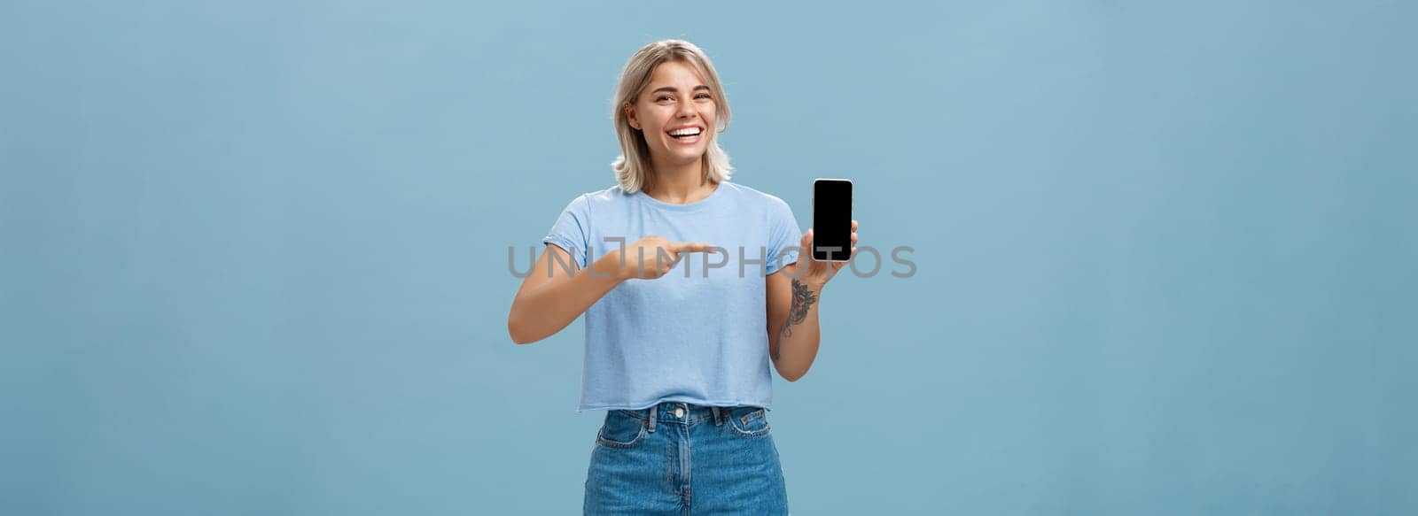 Look at this hilarious photo. Entertained attractive happy woman with fair hair in casual t-shirt and denim shorts showing smartphone at camera pointing at device screen smiling broadly over blue wall.