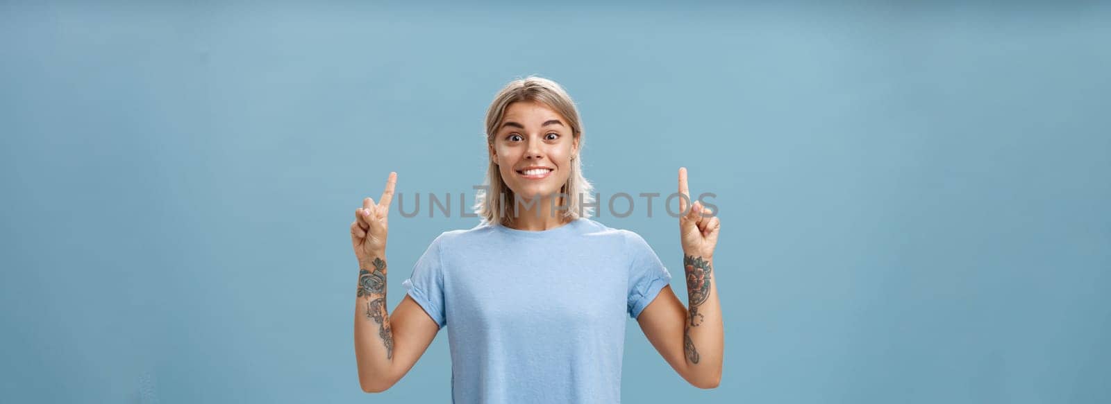 Lifestyle. Waist-up shot of excited and thrilled happy gorgeous blonde with short hairstyle and tattoos on arms smiling joyfully from amazement pointing up with raised index fingers posing over blue background.