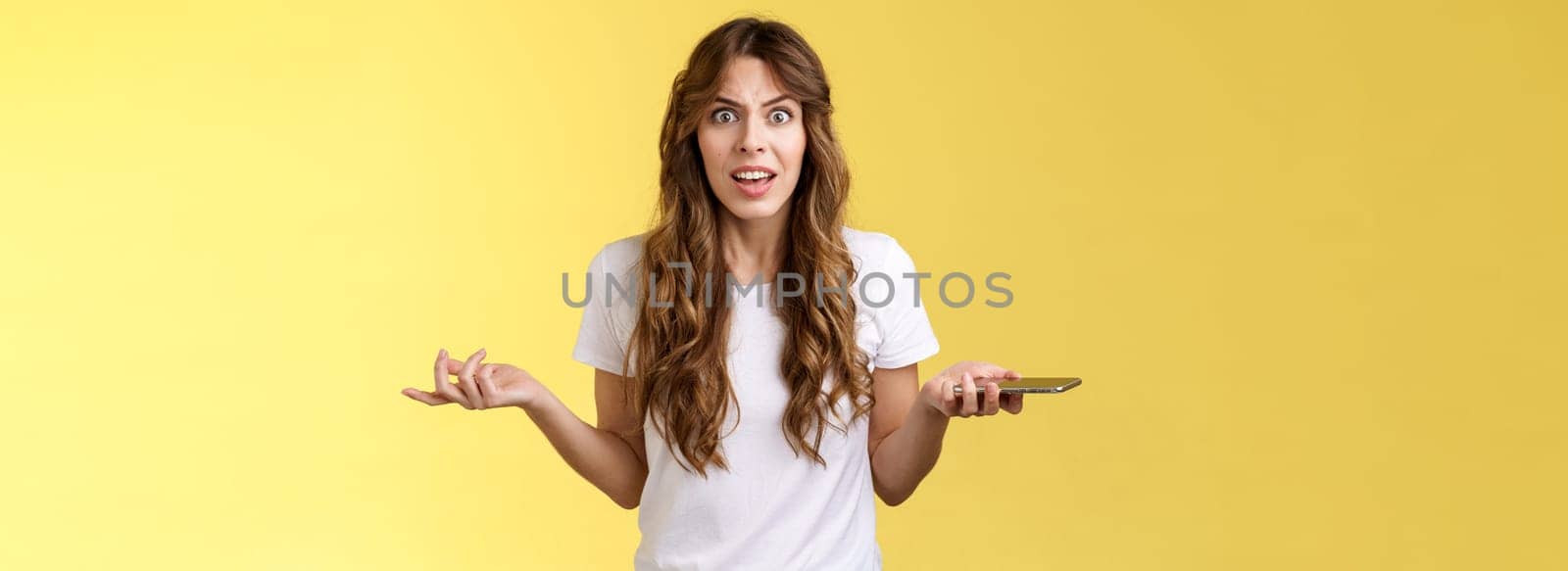 Pissed frustrated bothered curly-haired caucasian girl stare camera puzzled complain hold smartphone shrugging spread hands sideways dismay arguing after intense serious phonecall yellow background.