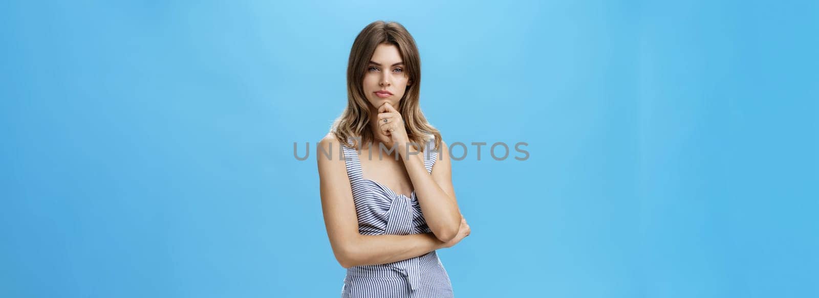 Girl using deductive method to solve mystery standing in thoughtful serious pose touching chin squinting seriously at camera and frowning thinking what decision make over blue background. People, gestures and emotions concept