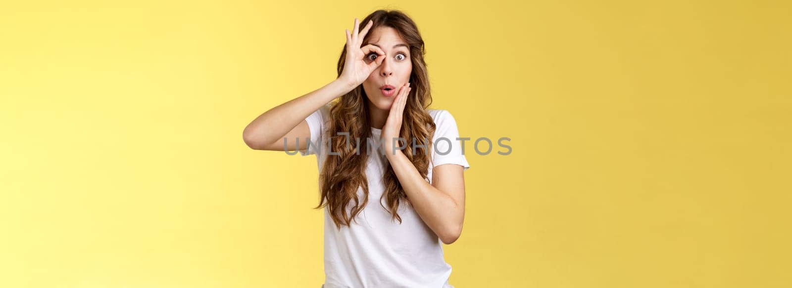 No way. Surprised impressed young funny attractive girl long chestnut hair fold lips admiration wow sound touch cheek astonished intrigued look through ok ring sign like monocle yellow background.