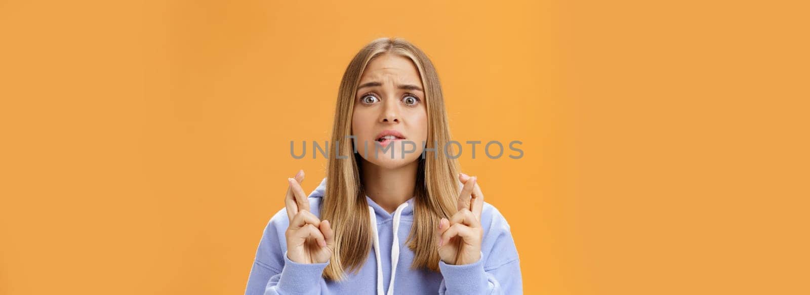 Worried anxious hopeful young woman with fair hair in stylish over-sized hoodie biting lower lip frowning concerned crossing fingers for good luck praying for dream come true against orange wall. Lifestyle.