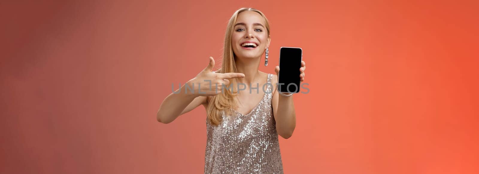 Boastful proud charming elegant blond woman in stylish evening dress show smartphone display proudly pointing mobile phone screen smiling showing photo boyfriend, standing red background.