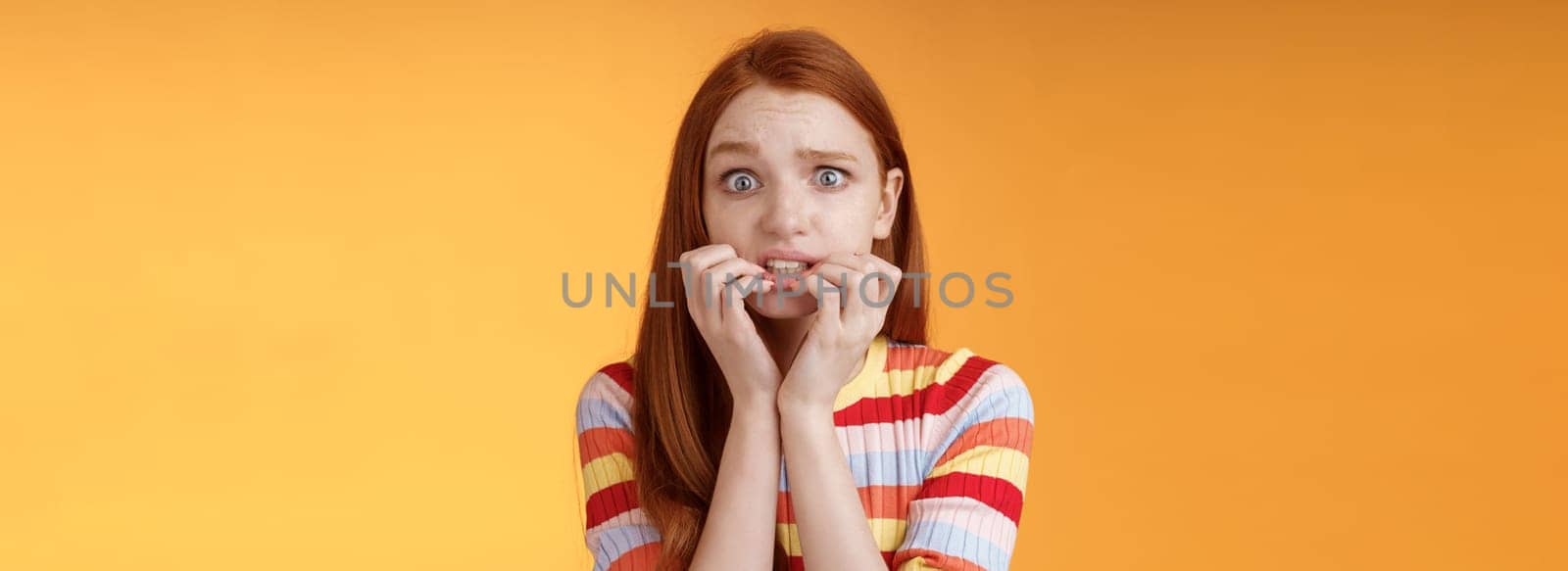 Anxious unconfident timid insecure redhead cute girl worry trembling fear consequences biting fingernails frowning grimacing frightened, standing nervously orange background awaiting scary moment.