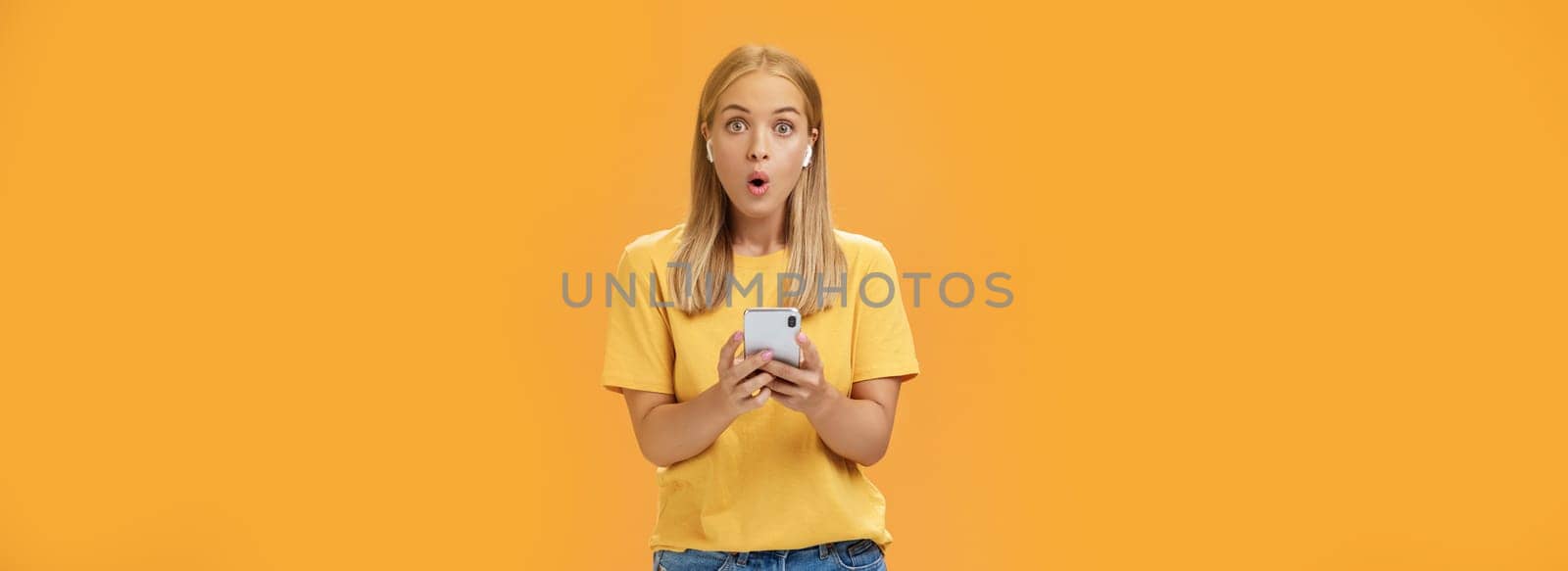 Woman surprised how awesome sounds new wireless earbuds holding smartphone wearing earphones folding lips in wow and amazement listening music impressed by good product over orange background by Benzoix