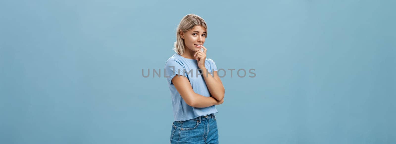 Studio shot of unconfident silly and insecure cute feminine girl with blond hair standing half-turned over blue background holding finger on lip while being shy ask question smiling hesitating by Benzoix