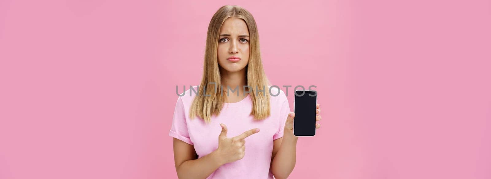 Gloomy and sad cute young female showing friend negative feedback about her project in internet pointing at smartphone screen standing concerned and upset against pink background. Technology and emotions concept
