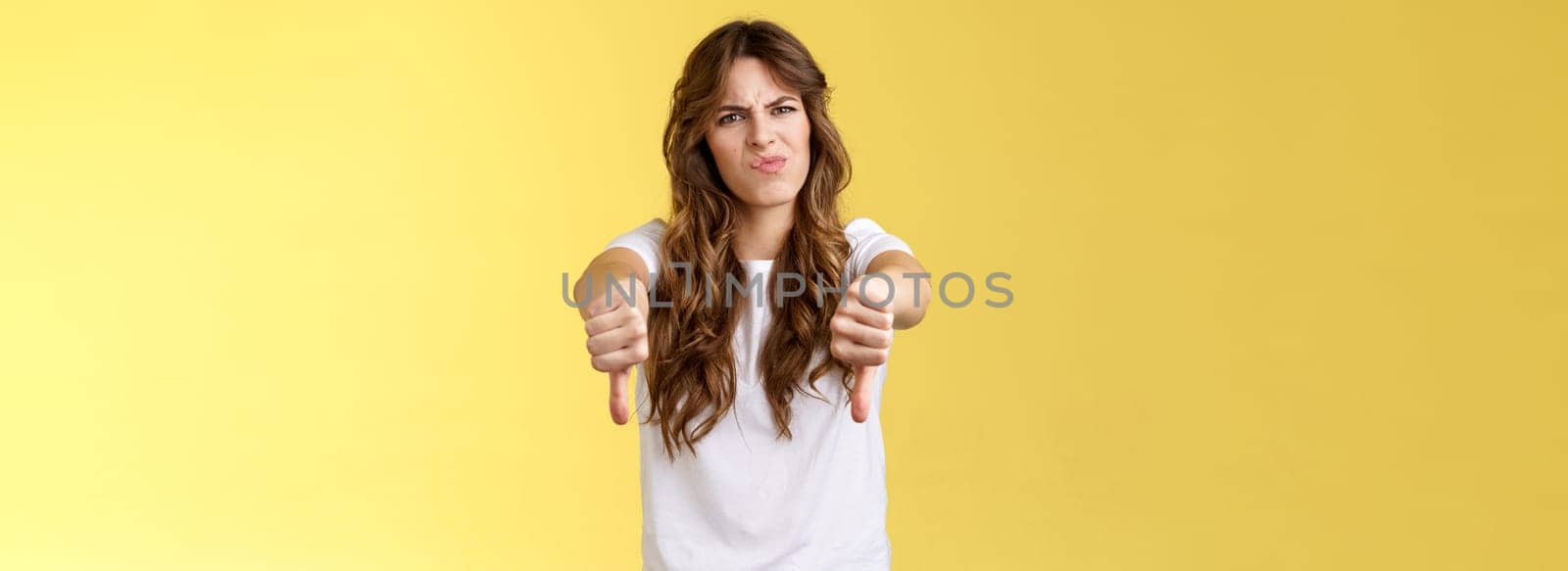 Lame bad idea disagree. Girl express strong dislike grimacing displeased frowning upset show thumbs down negative judgement disappointed awful movie stand yellow background unimpressed.