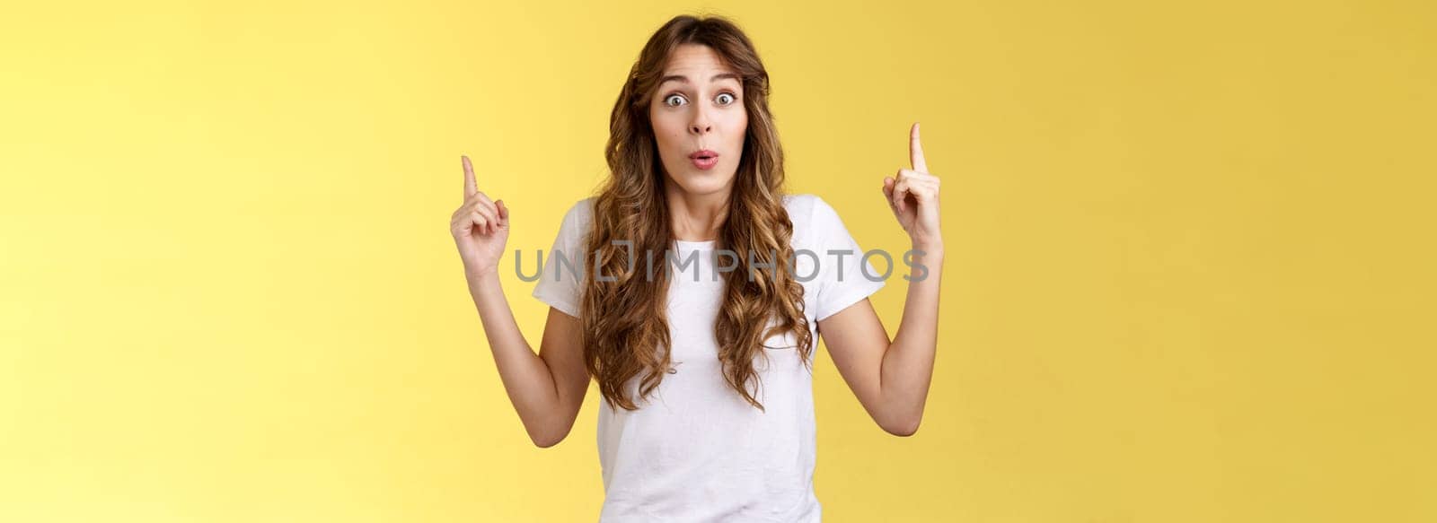 Impressed enthusiastic surprised ambushed cute female model fold lips wow admiration joy stare camera fascinated pointing up index fingers top copy space show you stunning offer yellow background. Lifestyle.