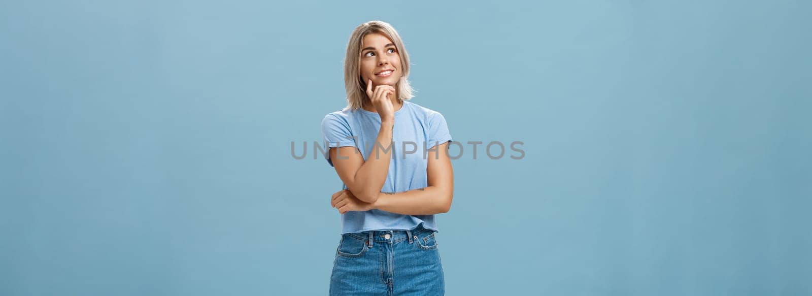 Dreamy thoughtful and creative artistic blonde woman in denim shorts and t-shirt smiling curiously touching lip and gazing at upper left corner feeling nostalgic, thinking over blue wall by Benzoix
