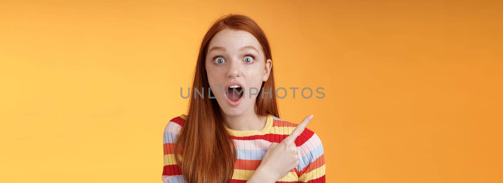 Impressed speechless young redhead 20s girl drop jaw gasping amused wide eyes staring camera surprised see awesome stunning new product pointing left index finger questioned, orange background.