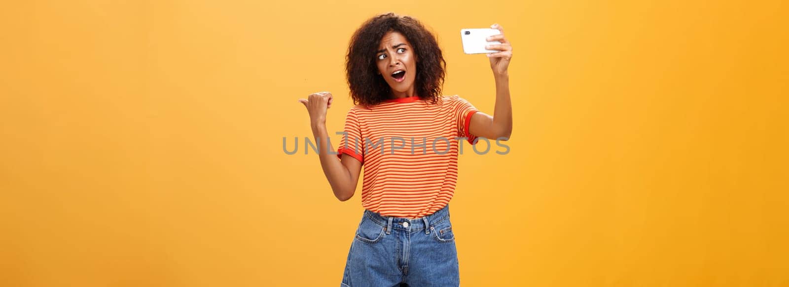Woman recording video blog pointing at strange object behind her. Portrait of concerned and curious stylish famous internet star holding smartphone talking in cellphone camera over orange background by Benzoix