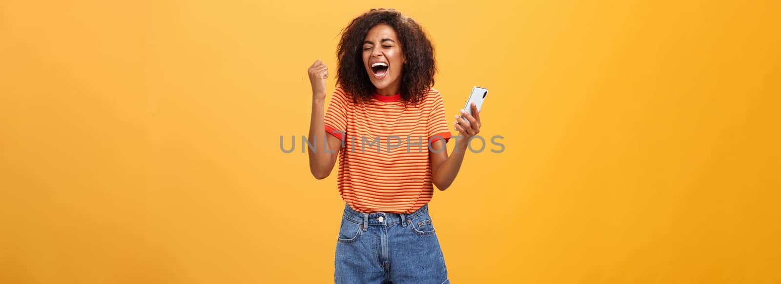 Portrait of ambitious happy young african american girl yelling from happiness and triumph clenching fist in joy and celebration feeling excited and relieved holding smartphone over orange wall. Lifestyle.