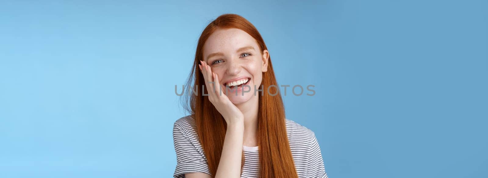 Charismatic talkative friendly-looking happy laughing redhead girl having fun discuss previous summer holidays make jokes chuckling touching face amused standing cheerful blue background.