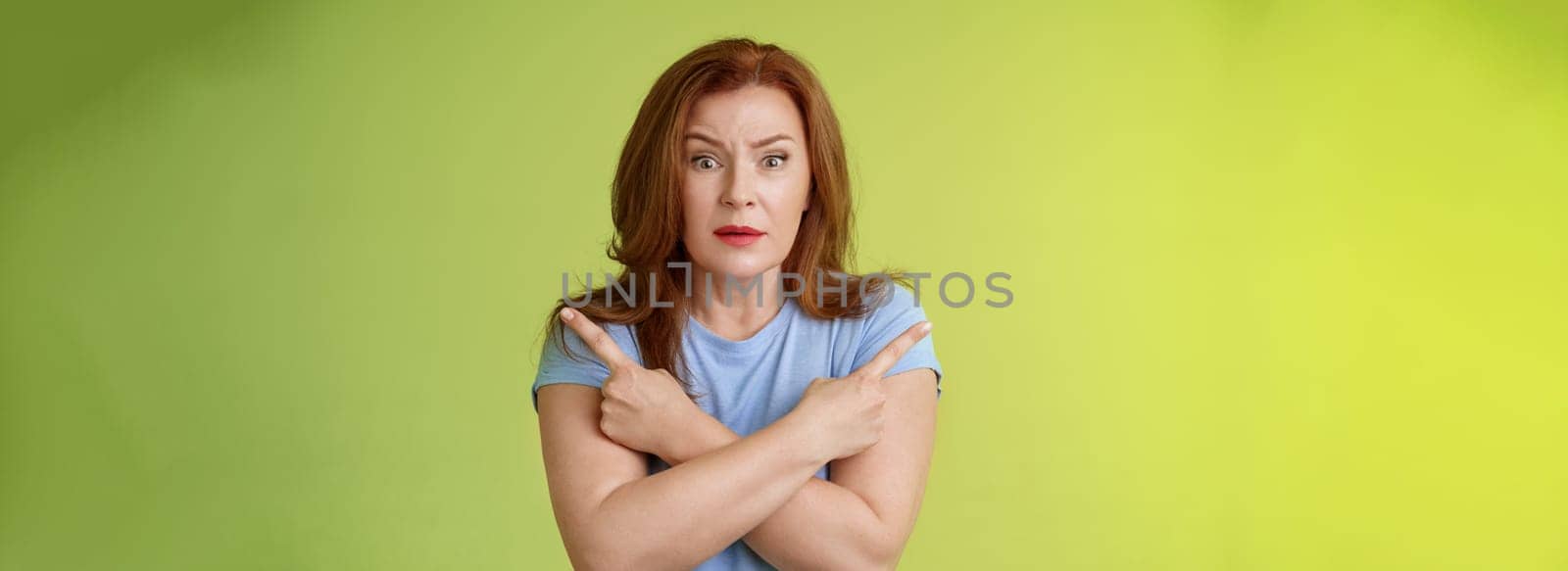 Help me choose. Worried unsure doubtful cute redhead middle-aged silly woman asking advice cross arms body pointing sideways left right look concerned camera make difficult decision green background.