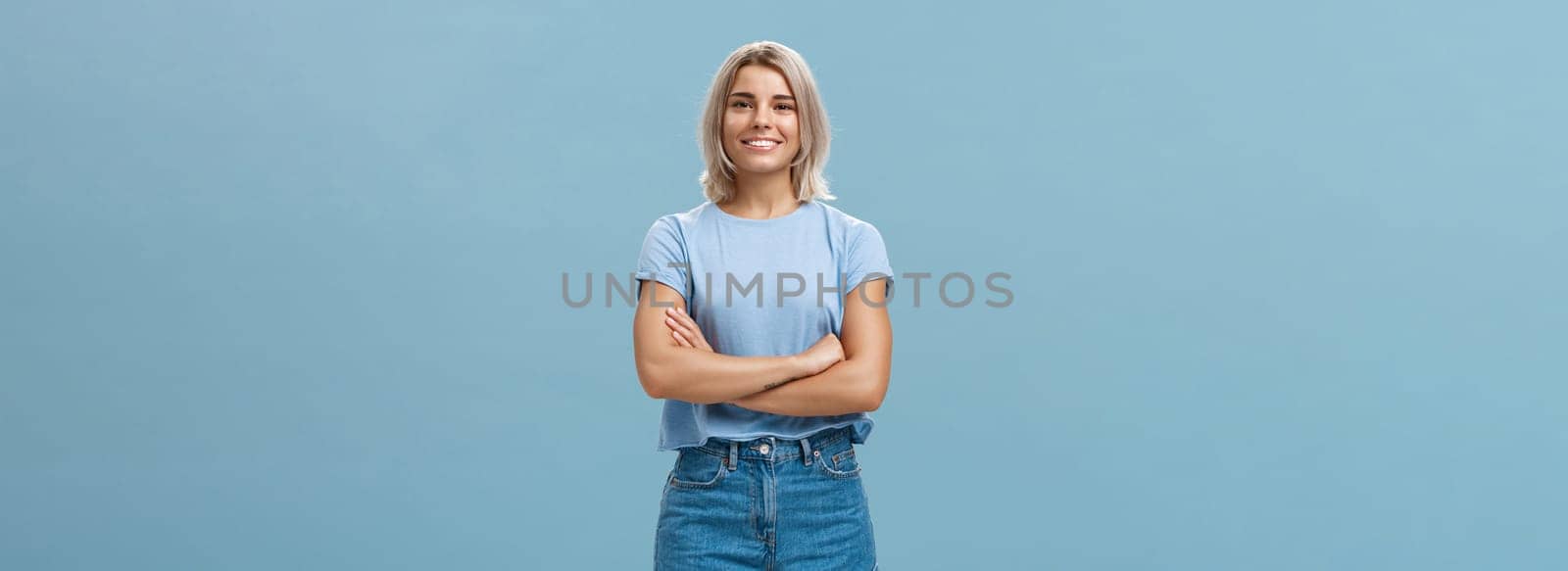 Ambitious good-looking young tanned female with blond hair standing in confident pose with hands crossed on chest smiling broadly standing in denim shorts over blue background. Copy space