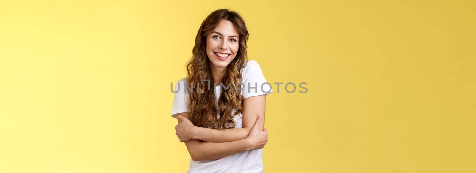 Tender lovely happy good-looking girl long curly hair hug herself wanna warm cuddles smiling toothy pleased friendly conversation having fun wear white t-shirt embracing own body yellow background by Benzoix