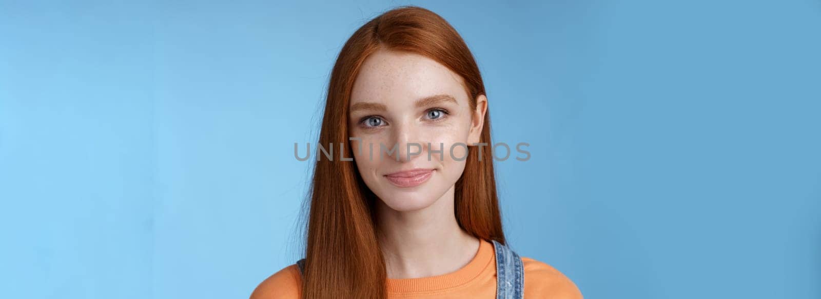 Outgoing young redhead girl blue eyes wearing orange t-shirt overalls smiling pleasantly casually talking standing good mood joyful emotions blue background, listening interesting conversation.