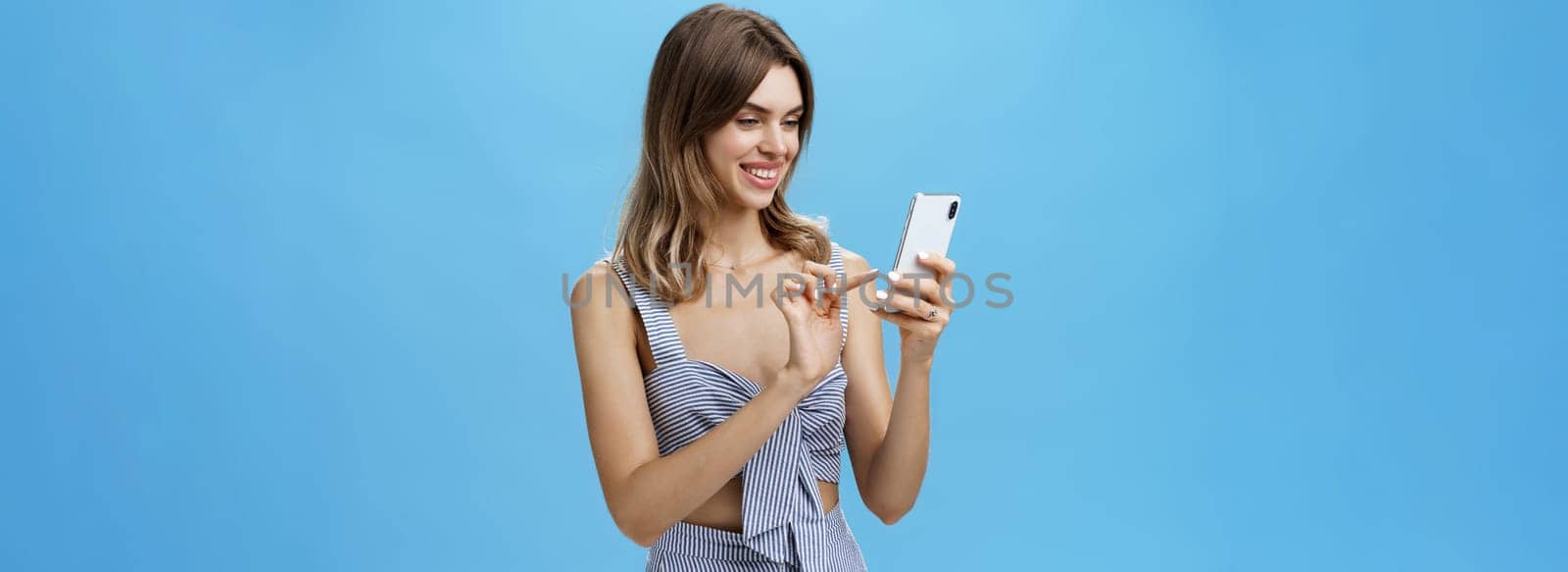 Portrait of stylish sociable good-looking female entrepreneur making appointment via smartphone holding cellphone typing message gazing at device screen with pleased carefree smile over blue wall. Technology concept