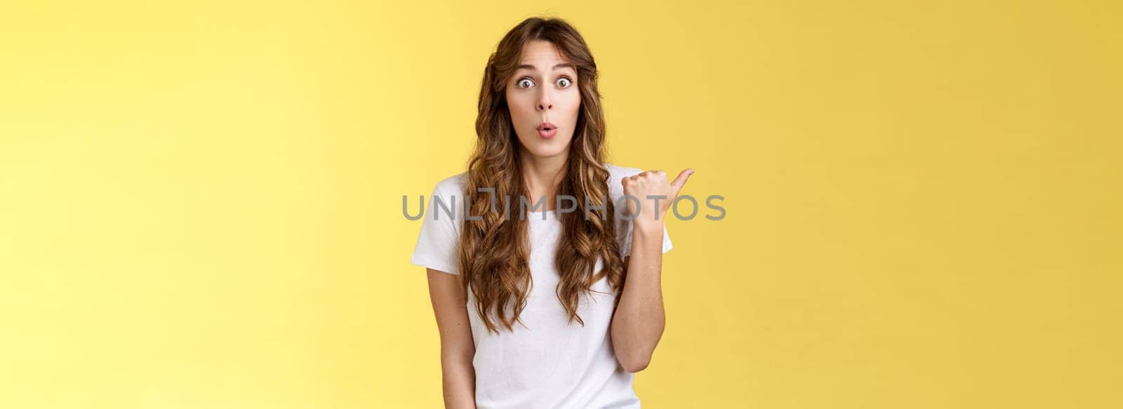 Wow interesting. Impressed surprised cute charismatic european girl curly haircut folding lips admiration fascintated pointing thumb left stare camera curious discuss gorgeous outfit coworker wear.