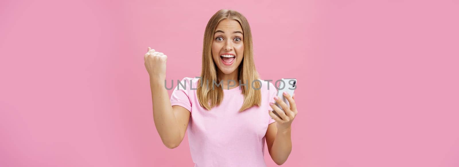 Girl beat own record in smartphone game raising clenched fist in cheer and triumph holding cellphone, smiling excited and happy at camera celebrating victory with joyful gesture over pink background. Technology and advertising concept