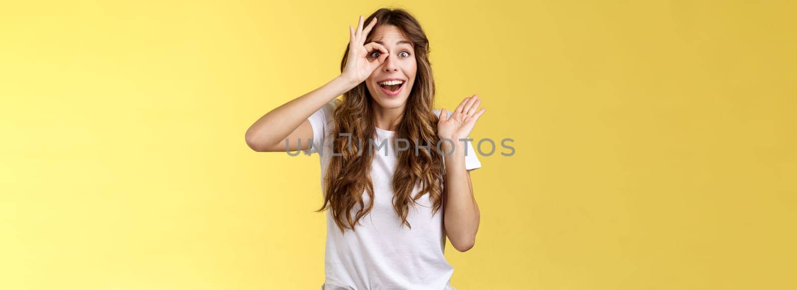 Oh hi I see you. Fascinated cute charming young girl waving palm say hello impressed look through okay ring gesture impressed finally fixed sight stand astonished amazed yellow background by Benzoix
