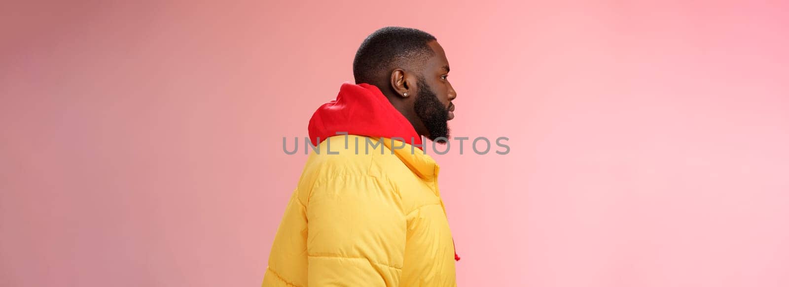 Profile studio shot bearded young 25s african guy in yellow jacket red hoodie look left normal unbothered relaxed expression standing queue order fastfood pink background, waiting take-away.
