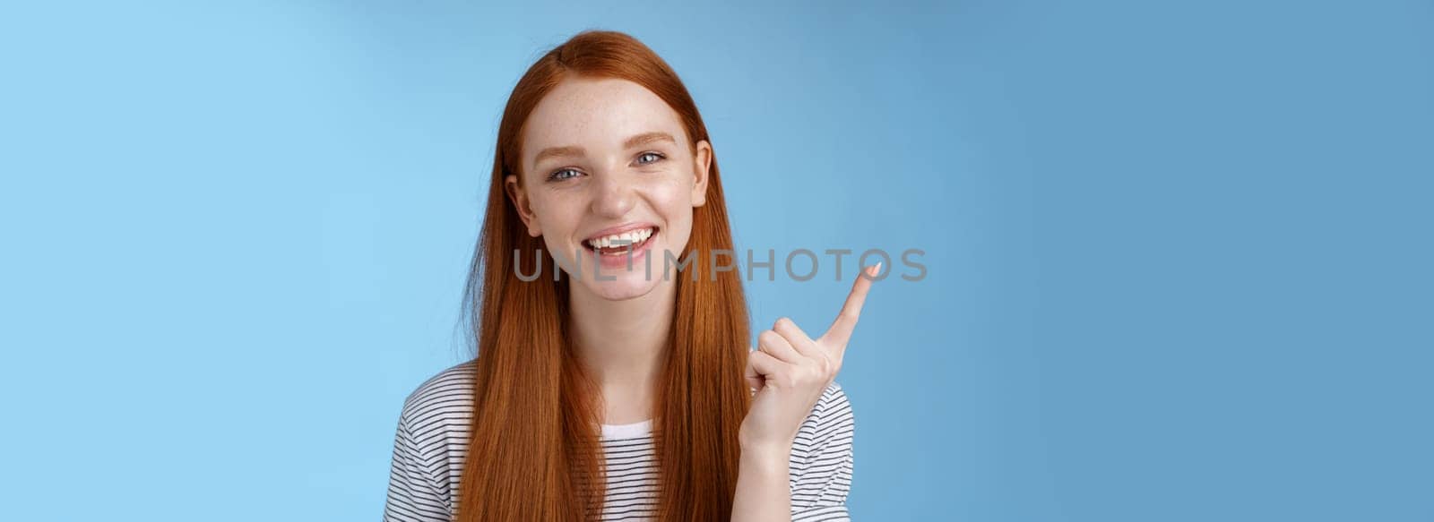 Happy charismatic redhead laughing young girl having fun looking carefree talking discussing new product sale pointing up index finger showing copy space advertisement, standing blue background.