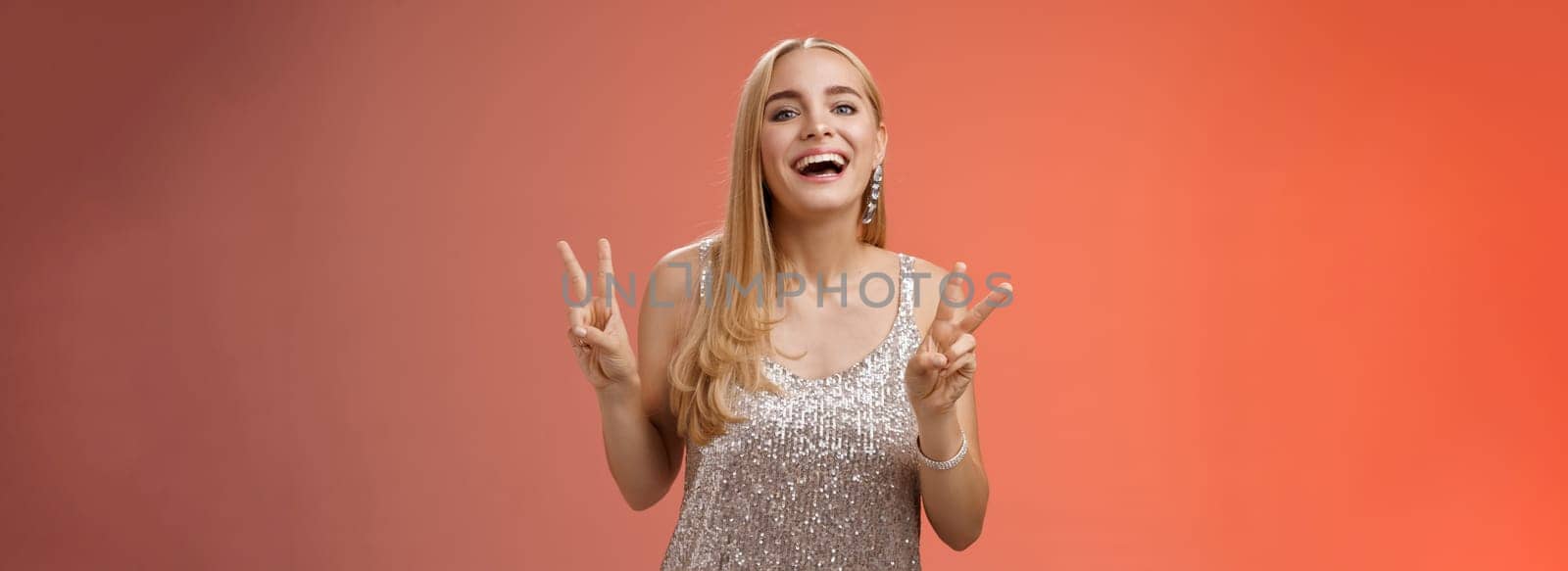 Funny carefree charming caucasian blond female model in silver stylish glamour dress show peace victory gesture smiling broadly have fun celebrating birthday party, standing red background.