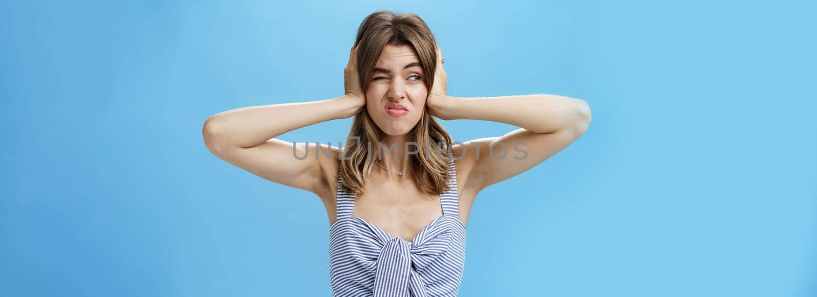 Girl feeling irritated by loud awful noise pressing hands to ears not hear annoying sound of drill smirking and frowning, squinting looking left displeased and disturbed, posing over blue background. Body language concept