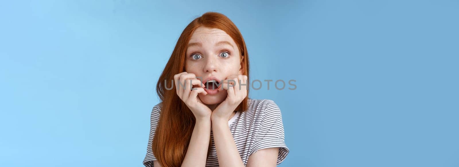Shocked speechless gasping young redhead girl staring impressed stunned watching important moment tv series biting fingers open mouth shook standing excited blue background anticipating.