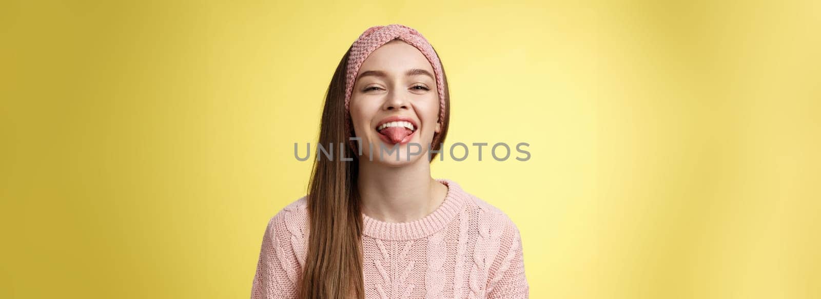 Positive entertained cute funny glamour young european girl in sweater, knitted trendy headband smiling fooling around showing tongue playfully, mocking friend enjoying sunny day over yellow wall.