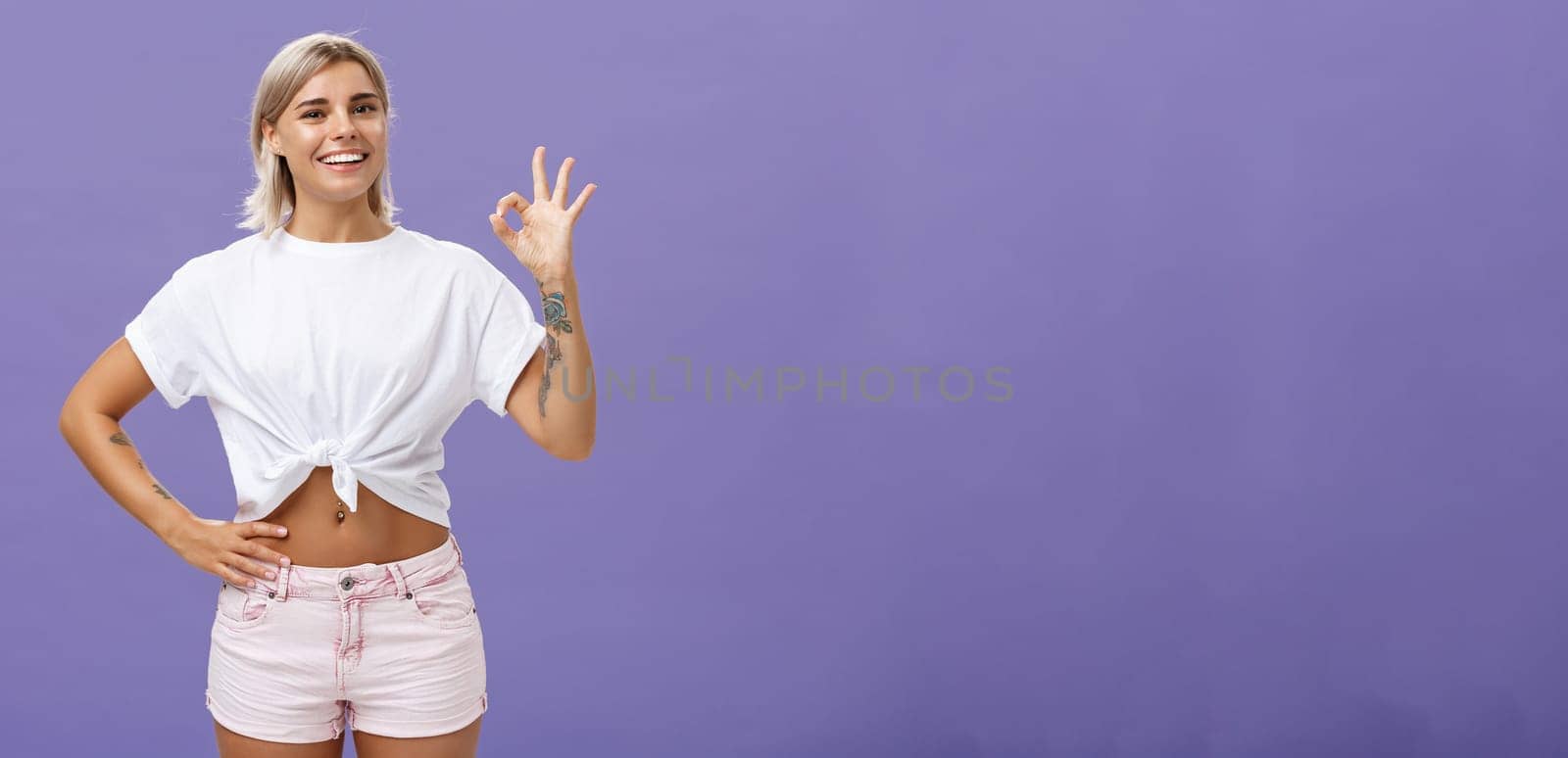 Excellent I like it. Portrait of satisfied good-looking happy girl with fair hair in white t-shirt and shorts showing ok or perfect gesture and smiling broadly holding hand on waist over purple wall.