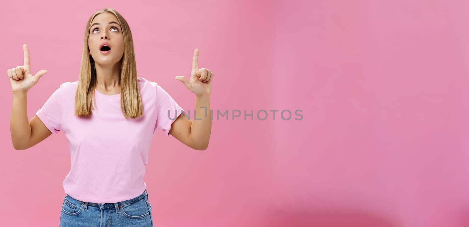 Portrait of amused and impressed curious attractive woman in t-shirt dropping jaw looking and pointing up astonished and intrigued watching interesting object in sky posing against pink background by Benzoix