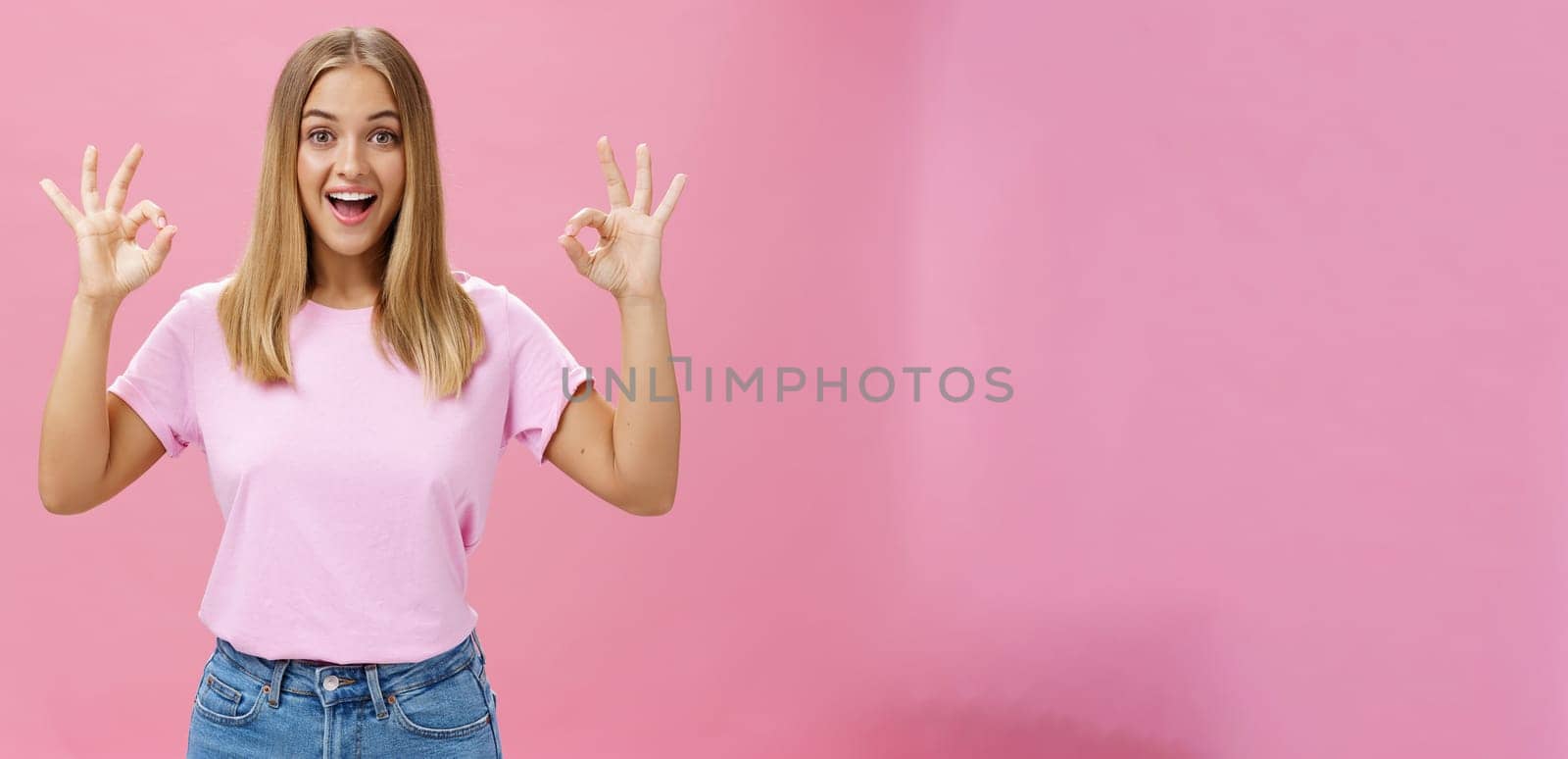 Portrait of enthusiastic attractive caucasian girl in trendy t-shirt and jeans showing okay or confirm gesture with amused broad smile standing pleased over pink background reacting to excellent news by Benzoix