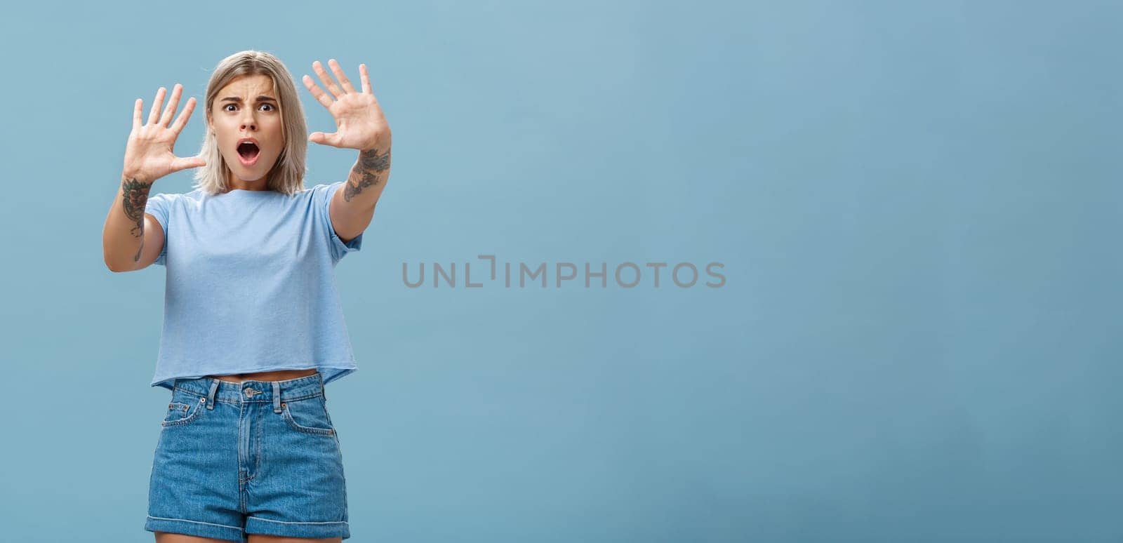 Woman terrified screaming and asking stop. Portrait of shocked panicking troubled blonde female in denim shorts and casual t-shirt pulling hands in no gesture dropping jaw and frowning. Copy space