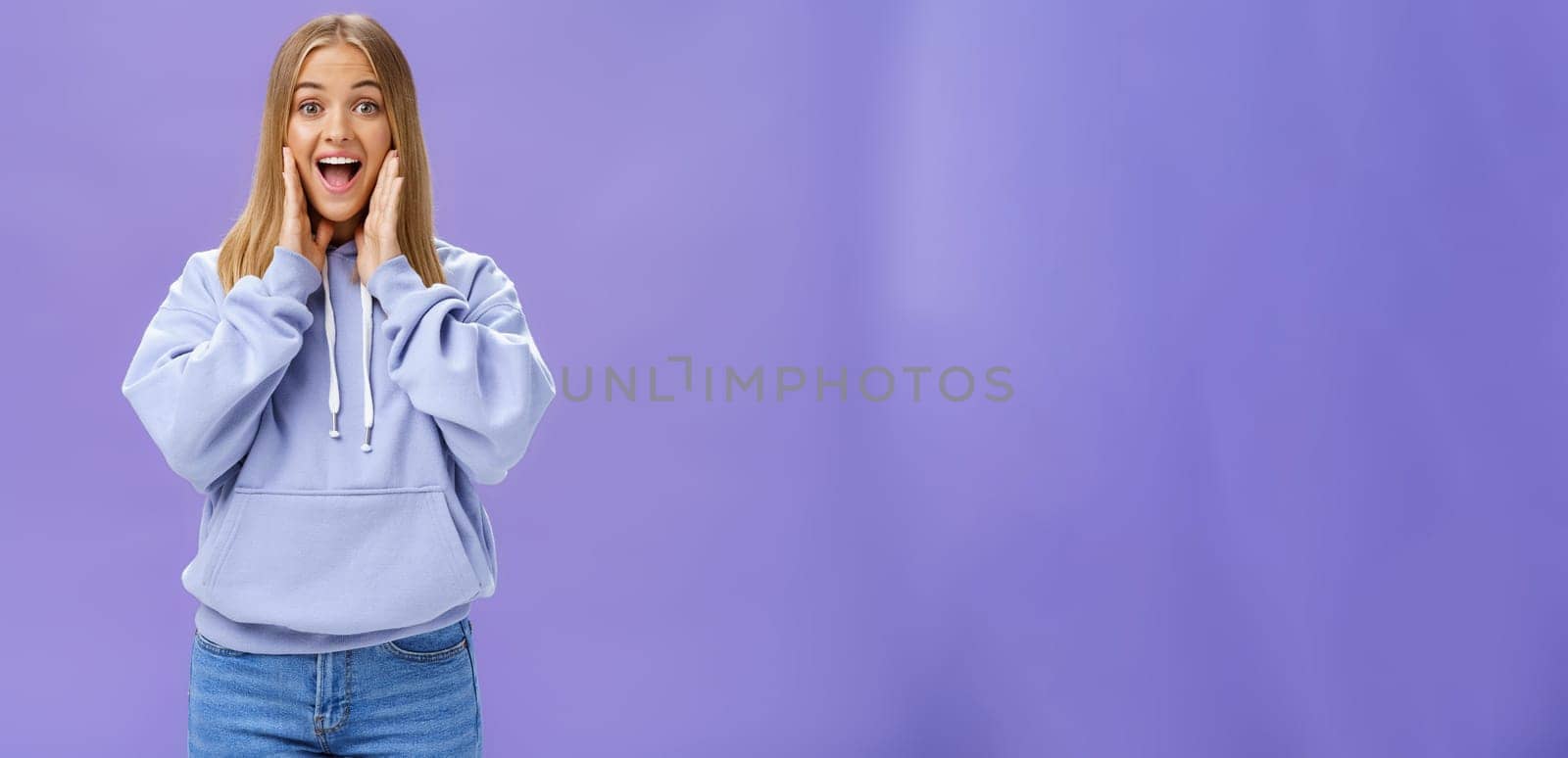 Woman joyfully smiling at camera and touching cheeks as if being surprised and happy see friend meeting person in airport feeling cheerful to reunite standing over purple background in cute hoodie. Emotions concept