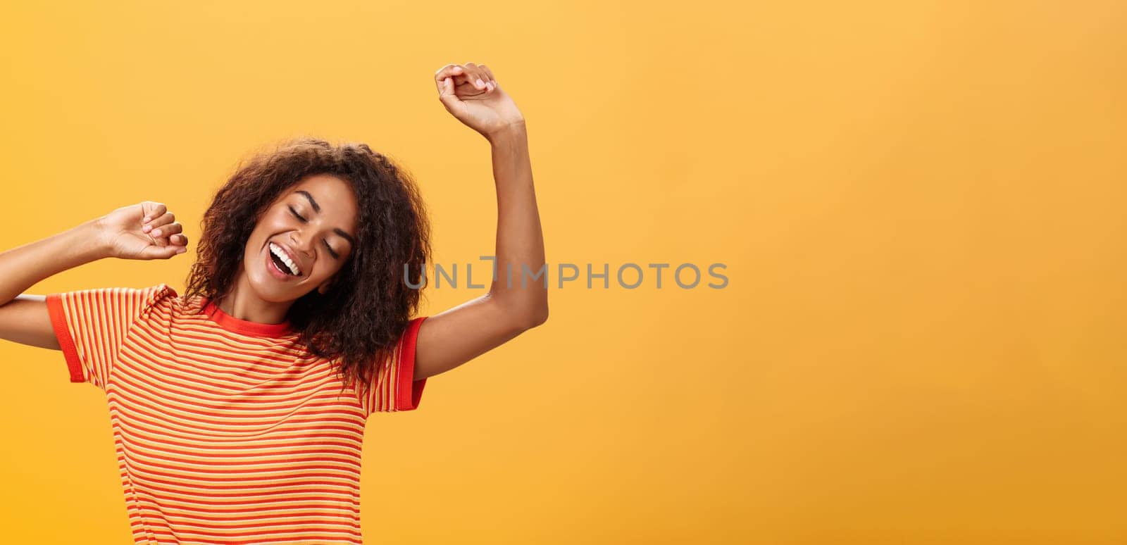 Waist-up shot of relaxed and relieved happy african american young female student in striped t-shirt stretching arms up closing eyes from delight and smiling having great nap over orange background by Benzoix