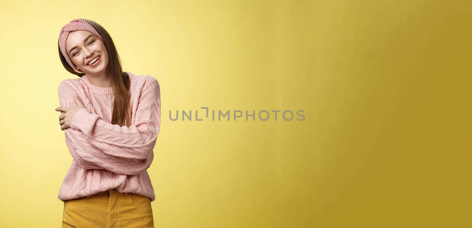 Cozy charming friendly young smiling girl in knitted warm comfortable sweater, grinning joyfully tilt head emracing herself, crossing arms, hugging feeling safe and happy, posing against yellow wall.