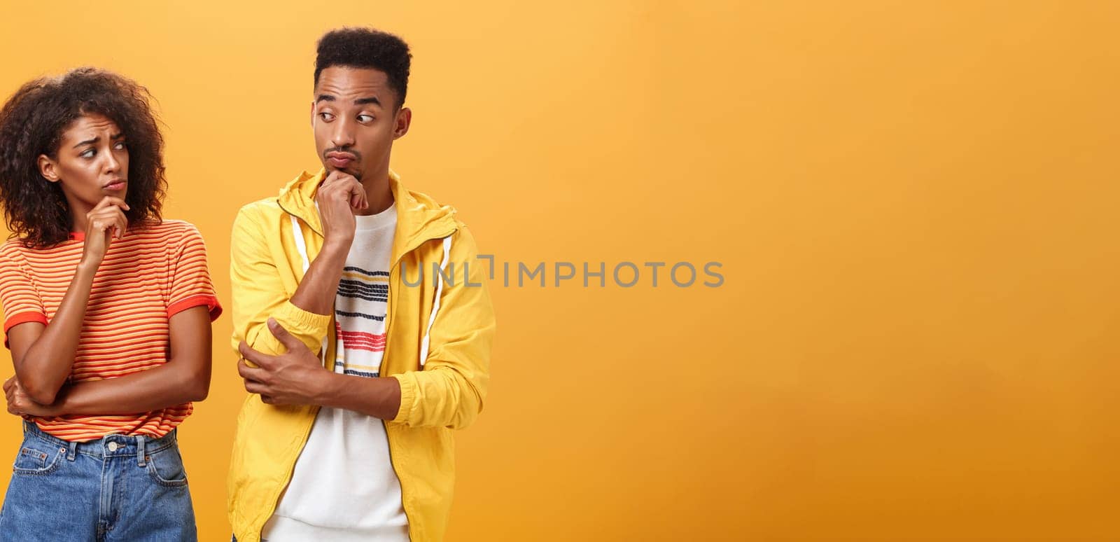 Silly confused girl standing gloomy and worried pursing lips and frowning holding hand on chin looking at boyfriend confused while guy glancing at girlfriend with curious expression over orange wall. Lifestyle.