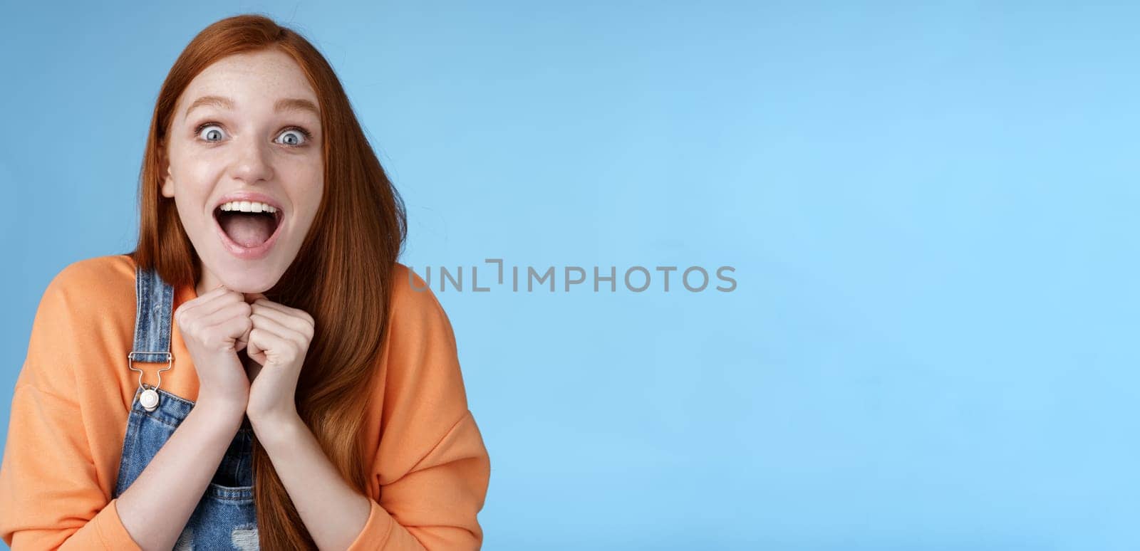 Cute redhead european girl blue eyes freckles reacting amused shocking rumor lift eyebrows drop jaw surprised smiling excited picked get role theatre play rejoicing astonished blue background by Benzoix